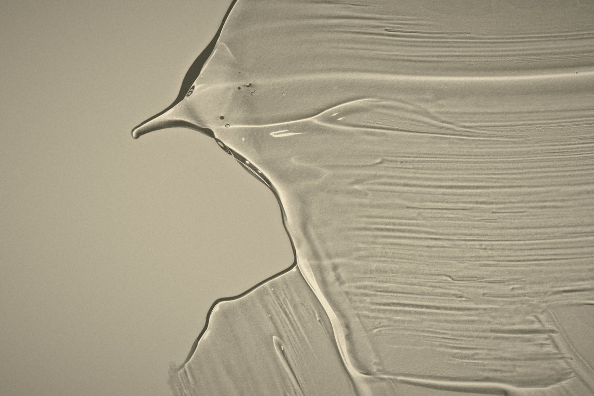 A close-up of a translucent, semi-opaque cosmetic or adhesive substance with a textured swipe pattern on a neutral beige background.